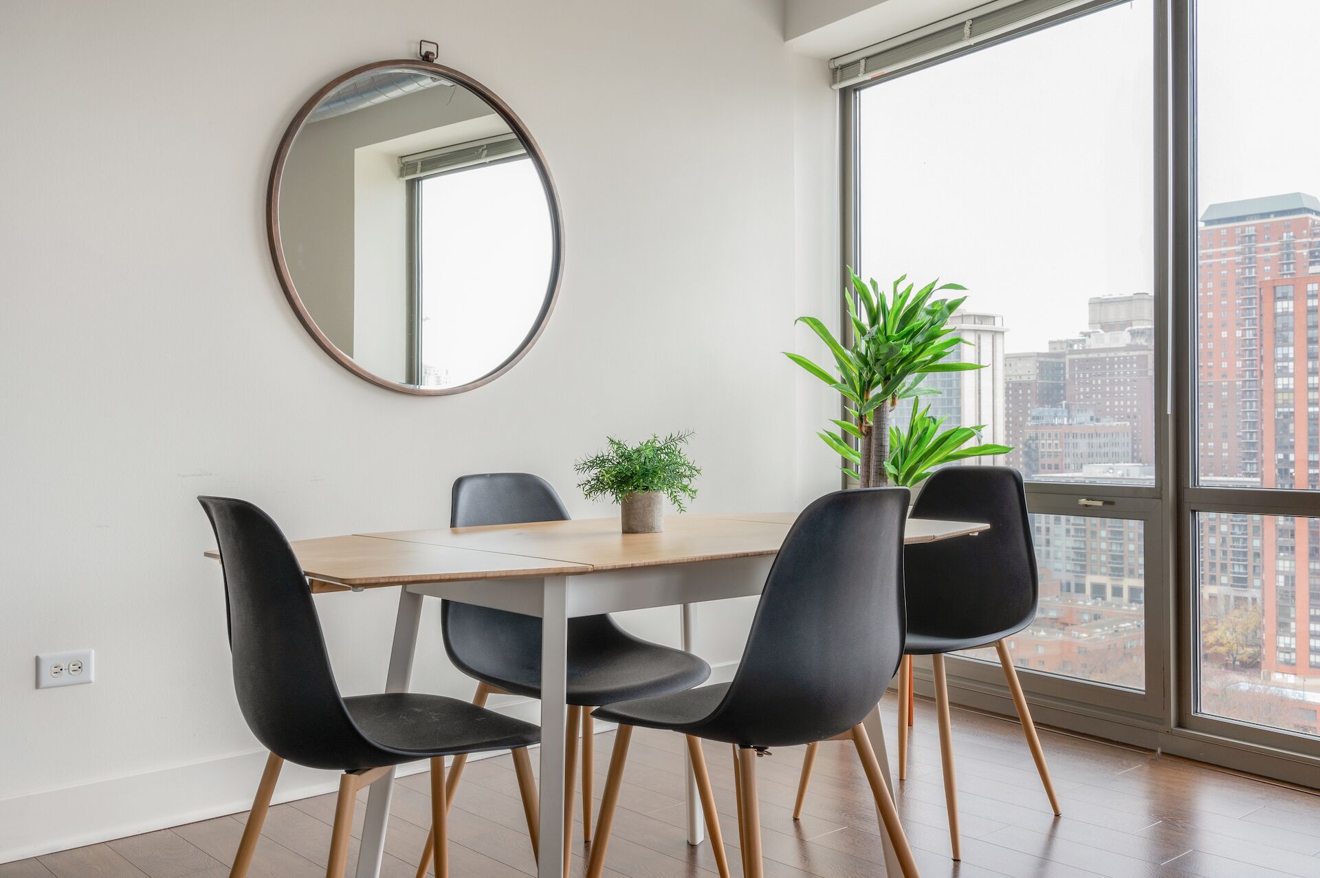 Un comedor con un espejo redondo, una mesa de madera, sillas negras y una planta en maceta.