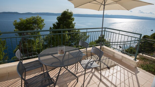 A terrace with a round table, chairs, and a large umbrella overlooking a body of water. - Apartments Andrea (Omis)