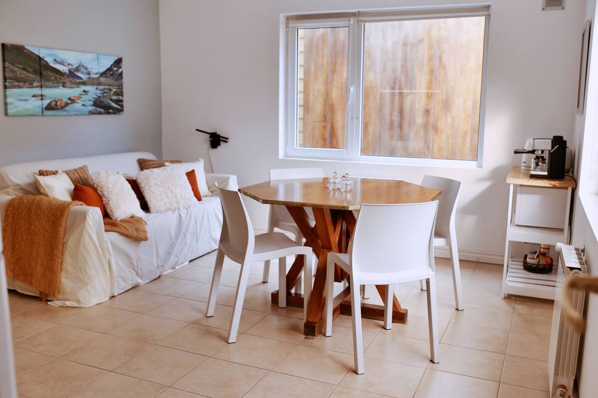 Een moderne woonkamer met een witte bank, een houten eettafel met stoelen, een groot raam en een koffieapparaat op een bijzettafeltje.