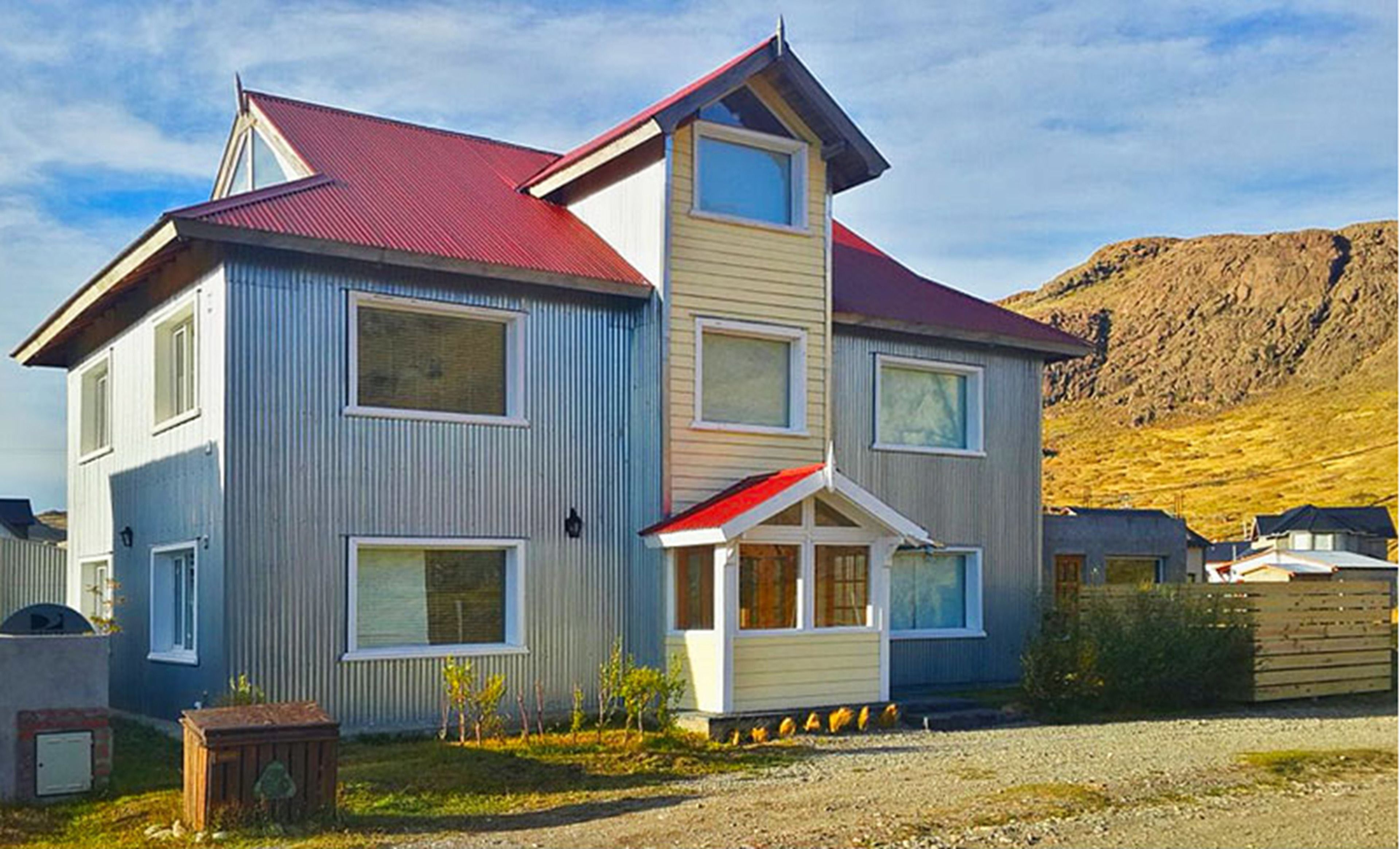 A two-story house with a red roof, a small porch, and a hillside in the background.