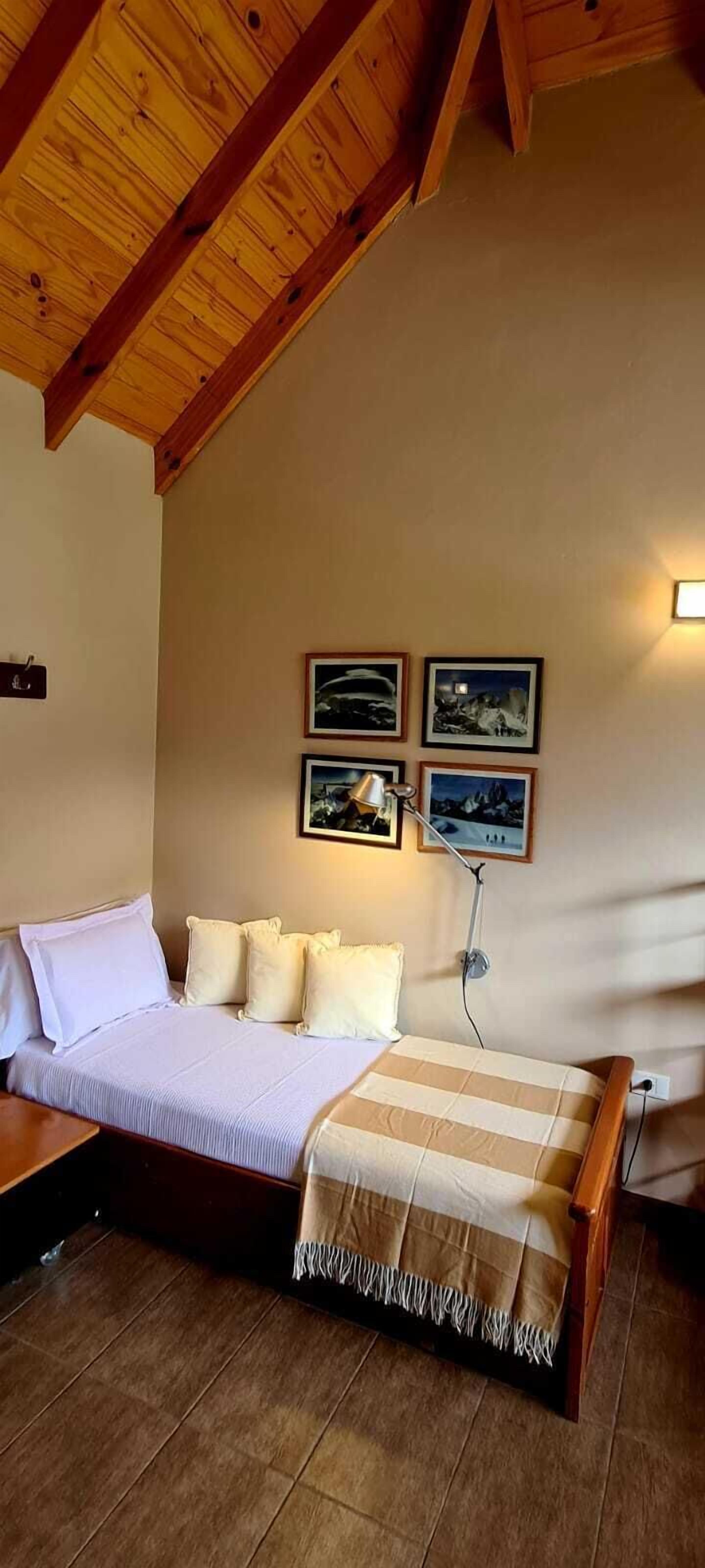 Un dormitorio con techo de madera, una cama con ropa blanca, una mesita de noche y una lámpara. Hay cuatro cuadros enmarcados en la pared.