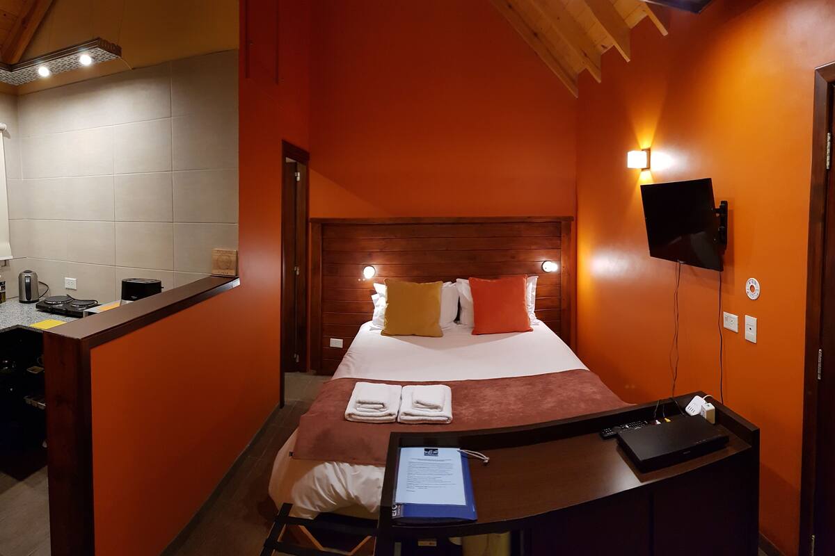 Een compacte hotelkamer met een bed, een bureau met een laptop, een aan de muur bevestigde televisie en een klein kitchenette.