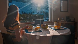 Uma pessoa sentada à mesa redonda com um laptop, uma tigela de frutas e um copo de suco de laranja. Uma janela mostra uma paisagem montanhosa com neve.