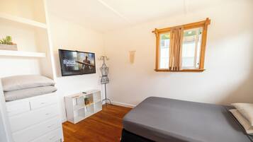 Un dormitorio con cama, televisor, armario, planta y ventana con cortinas.