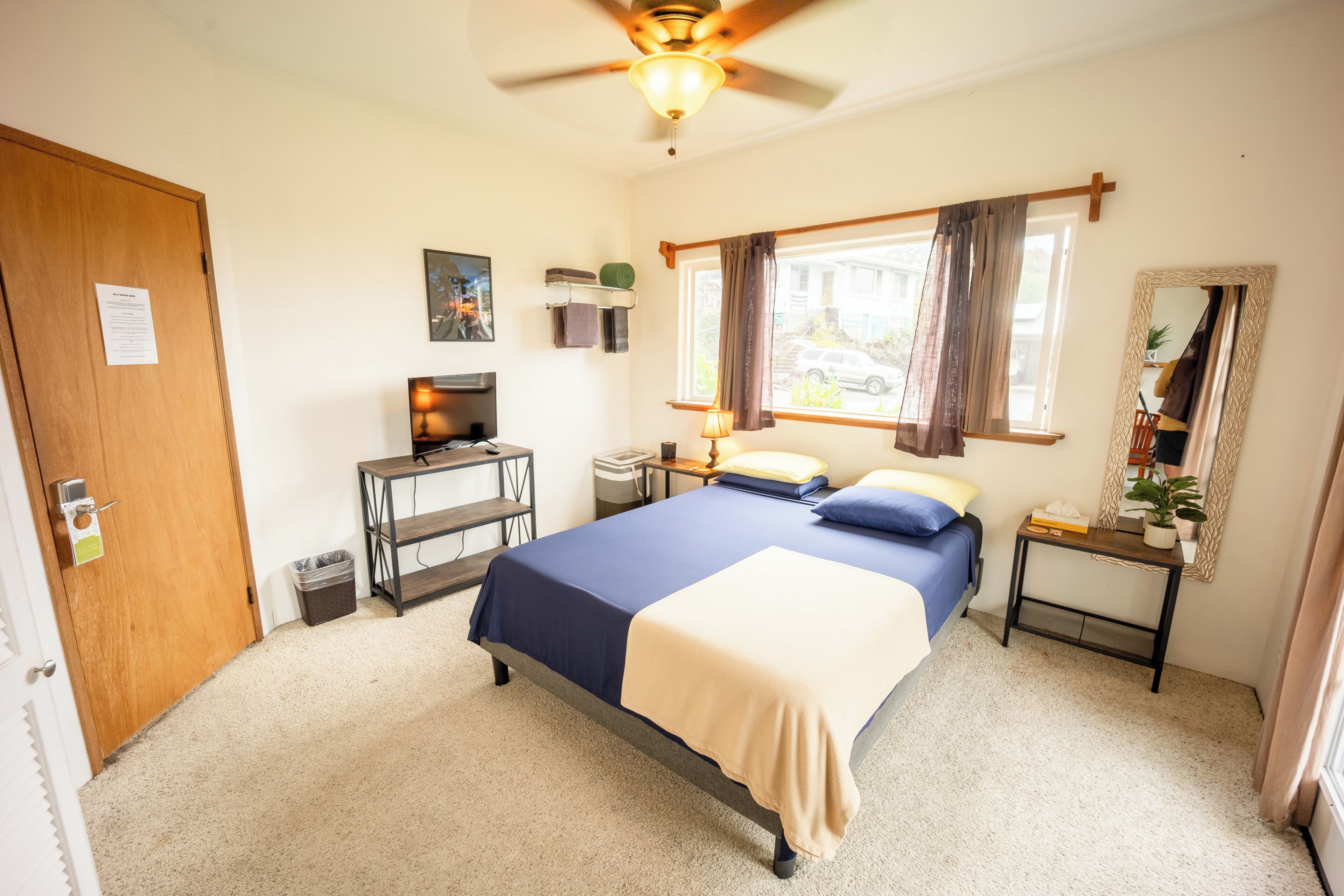 Ein Schlafzimmer mit einem Bett, einem Schreibtisch, einem Stuhl, einem Deckenventilator, einem Fenster mit Vorhängen und einem Spiegel an der Wand.