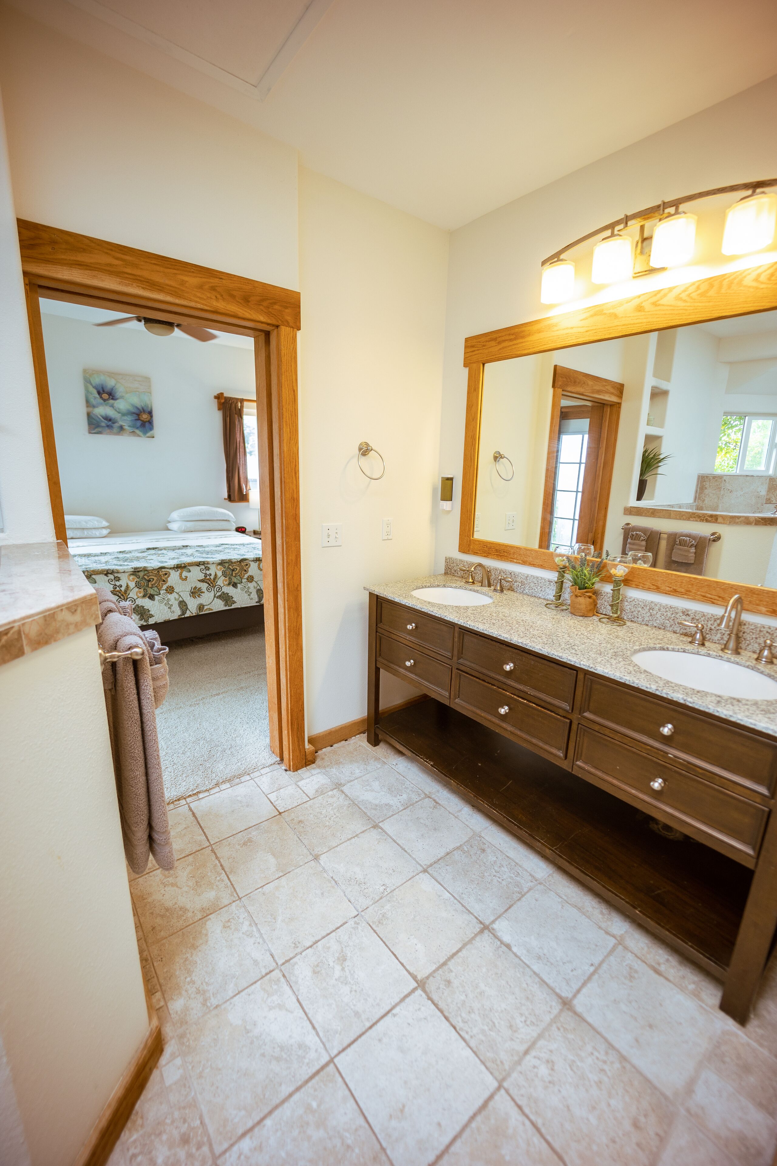 A bathroom with two sinks, a large mirror, and a bed visible through an open doorway.
