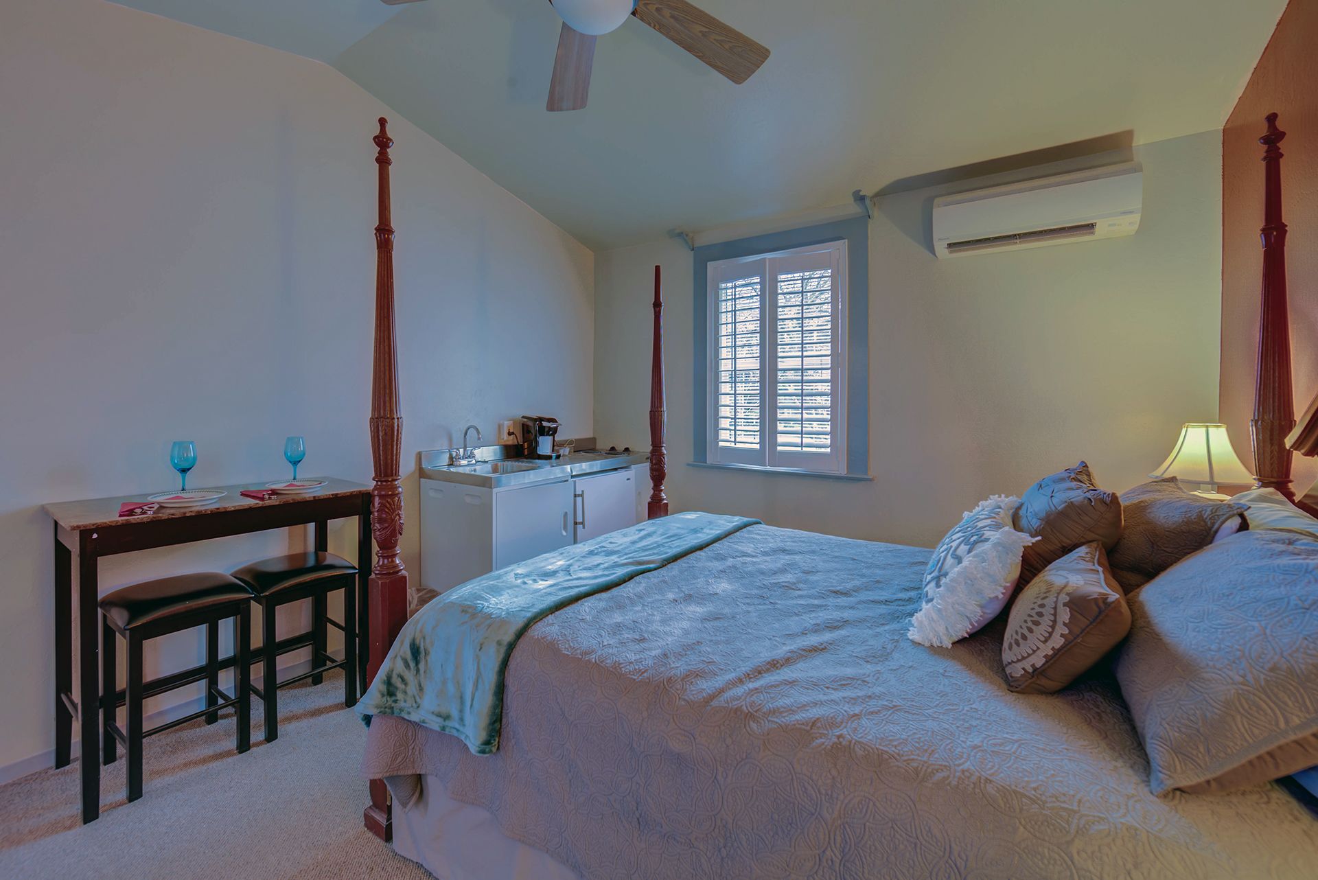 A bedroom with a bed, a desk, and a chair. There is a window with blinds, an air conditioning unit, and a ceiling fan.