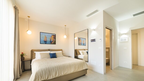 A modern hotel room with a large bed, bedside tables, a painting, a mirror, and a bathroom visible through an open door. - Garda Room (Peschiera del Garda)