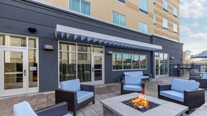 Un patio al aire libre con mobiliario de mimbre y una chimenea, junto a un edificio con ventanas amplias y un corredor cubierto.