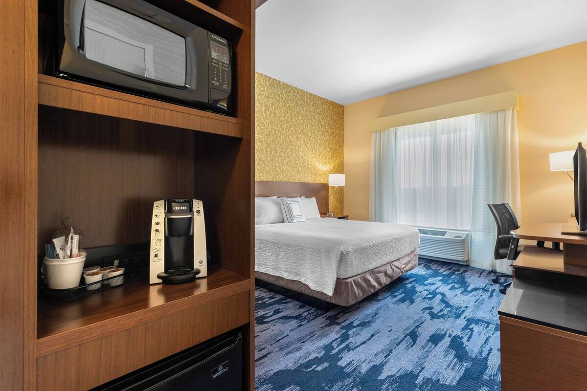 A hotel room with a bed, a desk, a microwave, a coffee maker, a TV, and a window with curtains.