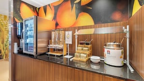 Una barra de desayuno con refrigerador, una exhibición de frutas y una máquina de café.