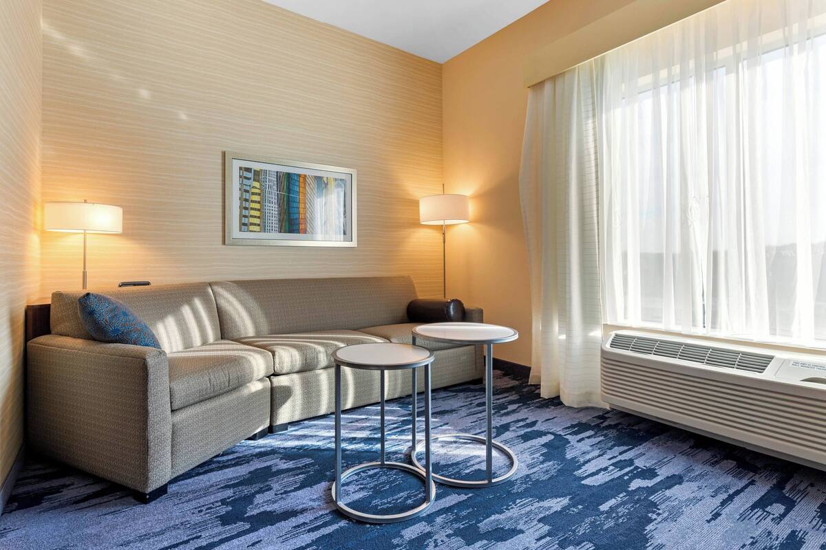 A hotel room with a sofa, two side tables, a framed artwork on the wall, and a large window with curtains.