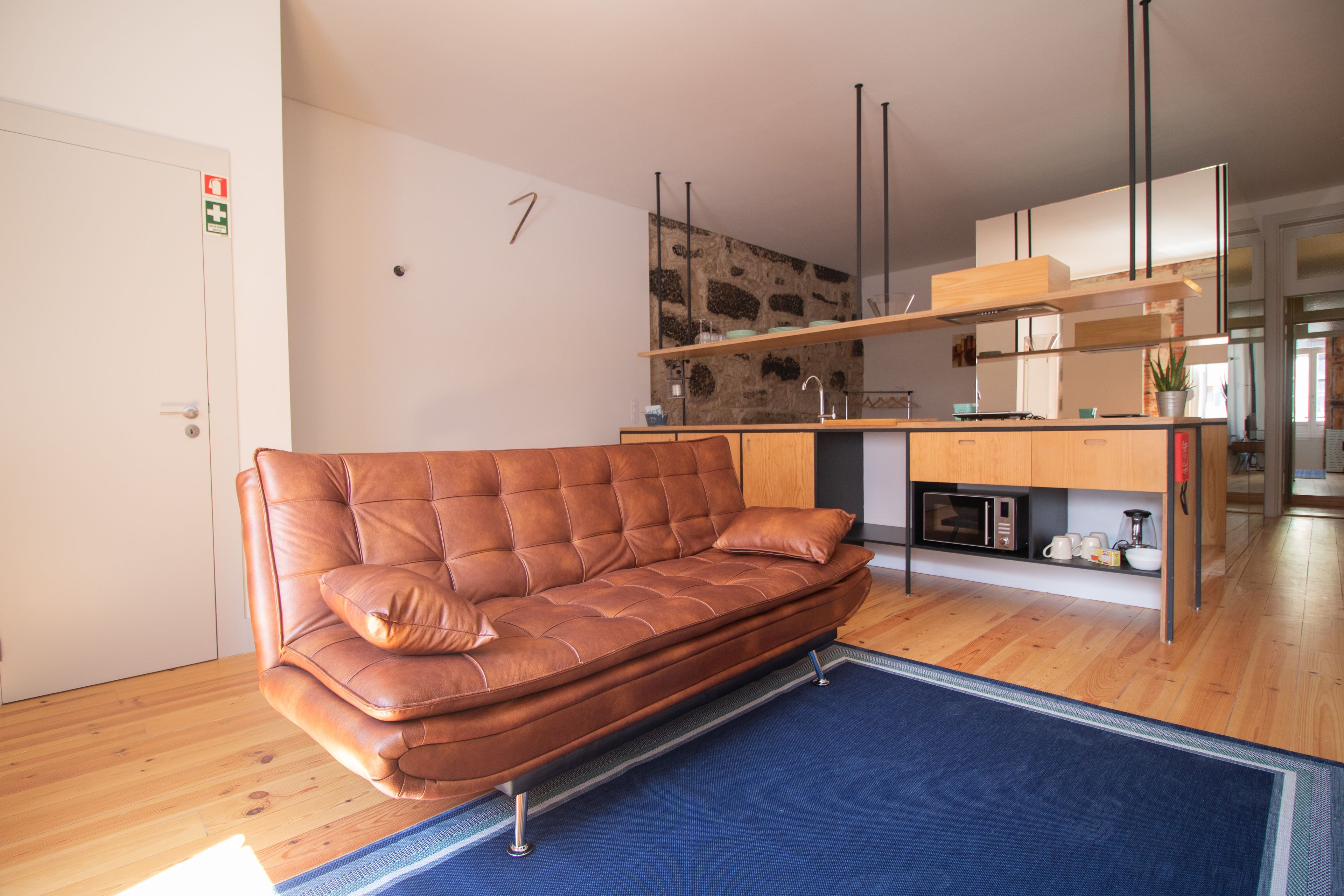 A modern living room with a brown leather sofa, a blue rug, wooden flooring, a kitchen area with wooden cabinets, and a stone accent wall.