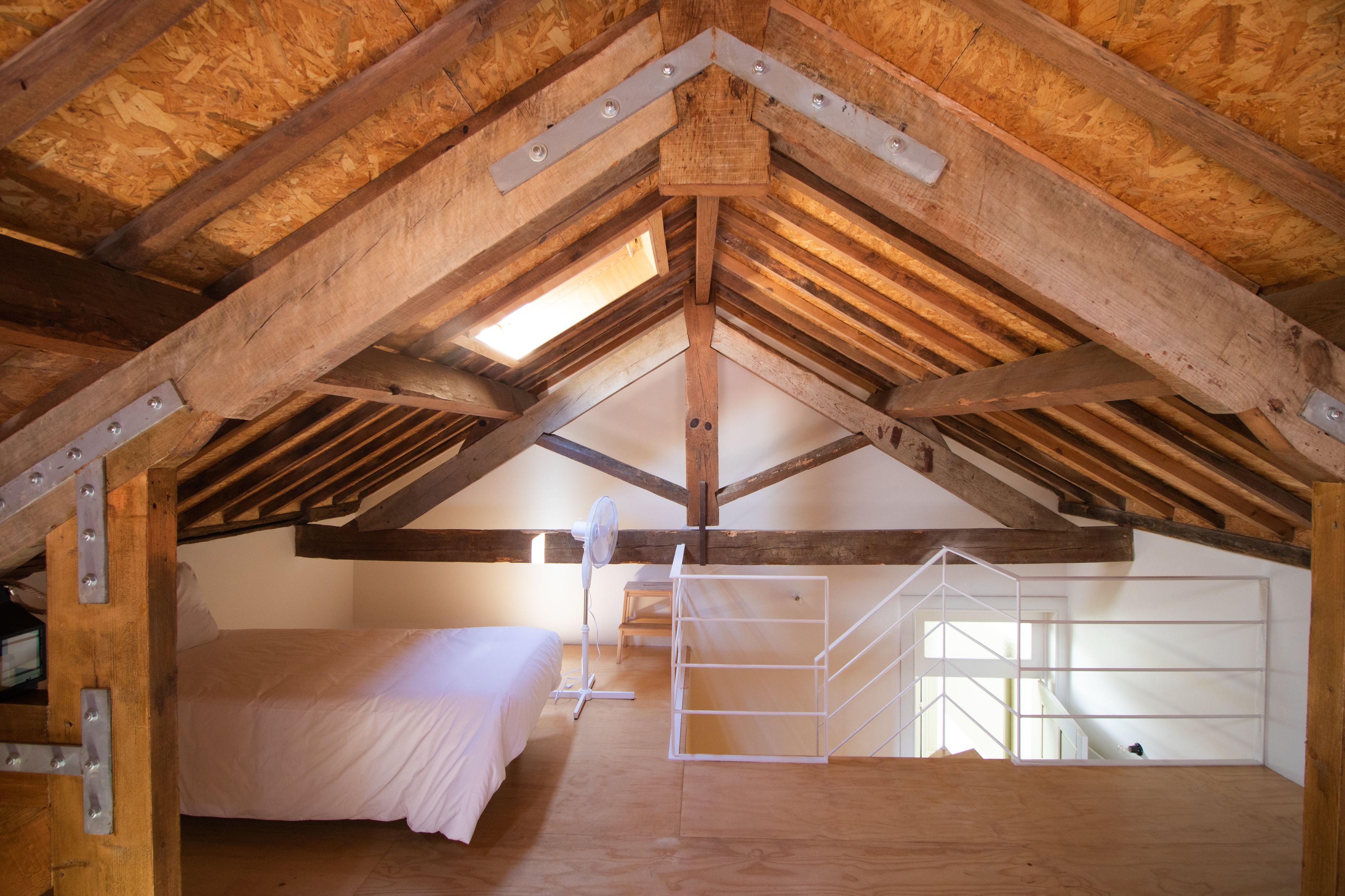Slaapkamer in loftstijl met zichtbare houten balken, een bed, een ventilator en een witte ladder.