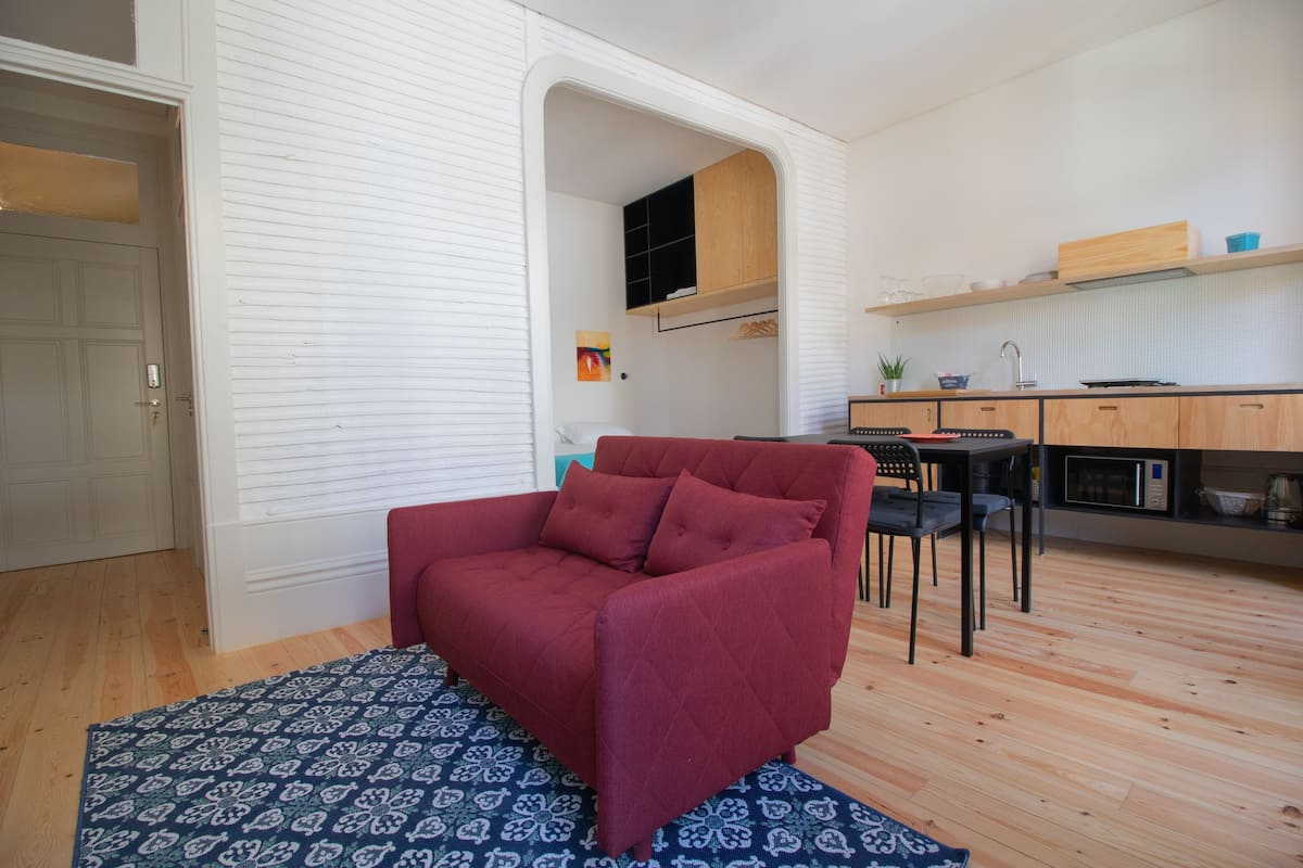A compact living space with a red sofa, patterned rug, wooden flooring, and a kitchenette area.