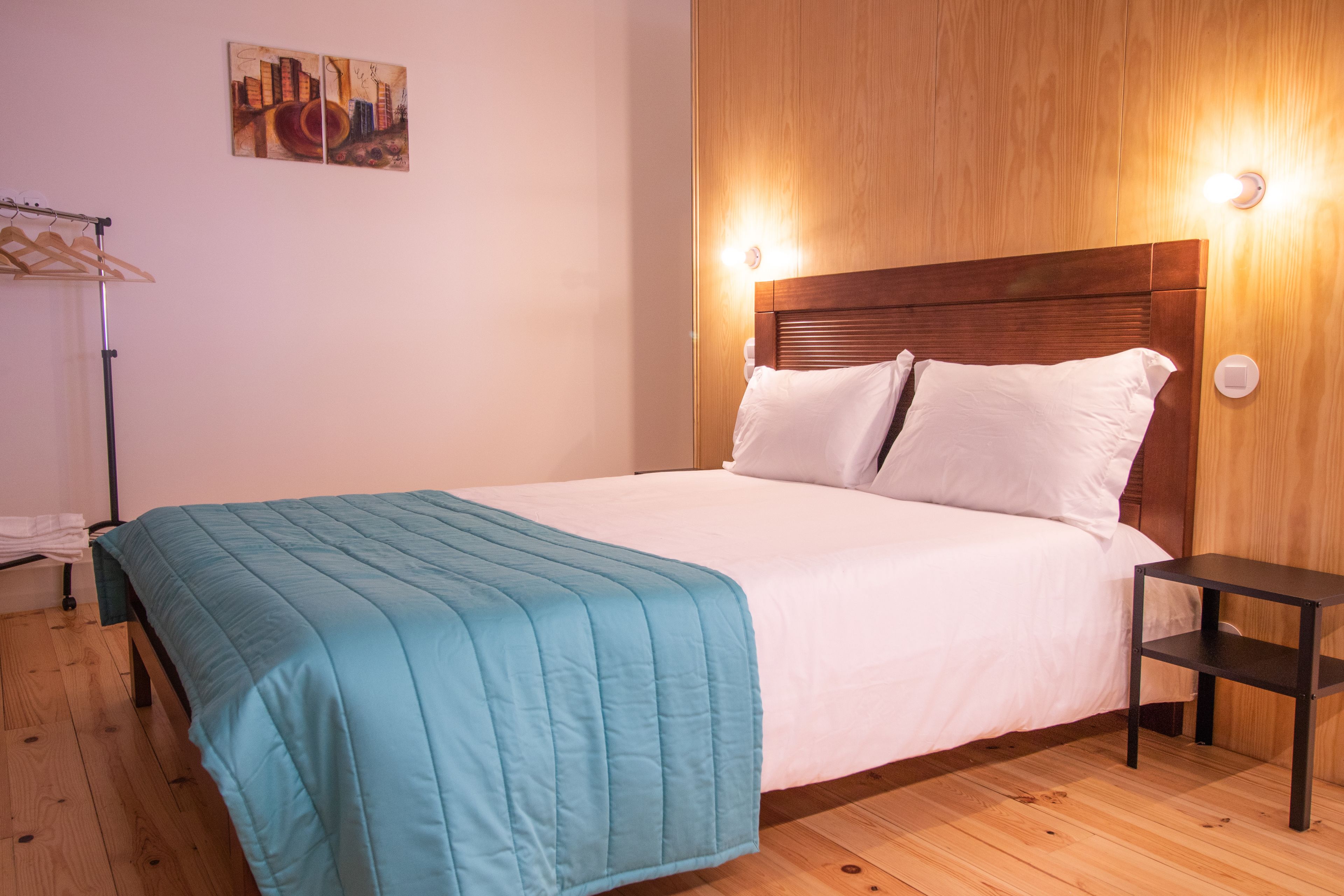 Una cama cuidadosamente hecha con una colcha color turquesa y almohadas blancas, un cabecero de madera, una mesita de noche y un aplique de pared.