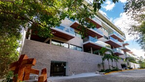 A modern building with multiple balconies, a brick facade, and a paved entrance area with a large sculpture. - Cruz con Mar by Playa Paradise (Playa del Carmen)