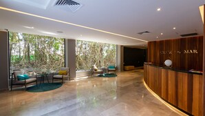 A modern hotel lobby with a wooden reception desk, seating area with chairs and a small table, large windows with a view of trees, and a polished marble floor. - Cruz con Mar by Playa Paradise (Playa del Carmen)