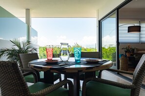 A round table with chairs, a pitcher, and two glasses on a balcony with a view of buildings and greenery.