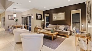 Een moderne woonkamer met een bank, salontafel en stoelen. Er is een eethoek met een tafel en stoelen. De kamer heeft een groot raam en een spiegel aan de muur.