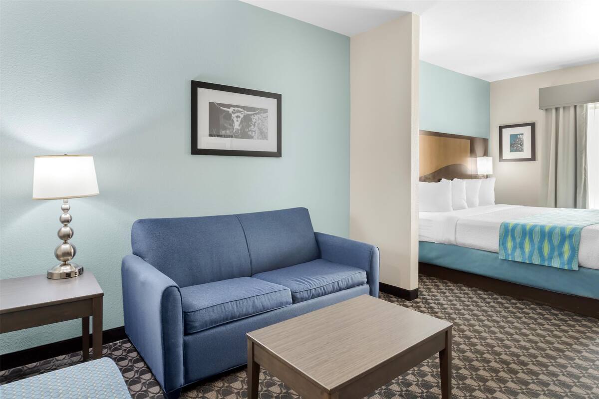 Una habitación de hotel con un sofá azul, una mesa de centro de madera, una alfombra estampada, una cama con colcha azul y blanca, y un cuadro enmarcado en la pared.