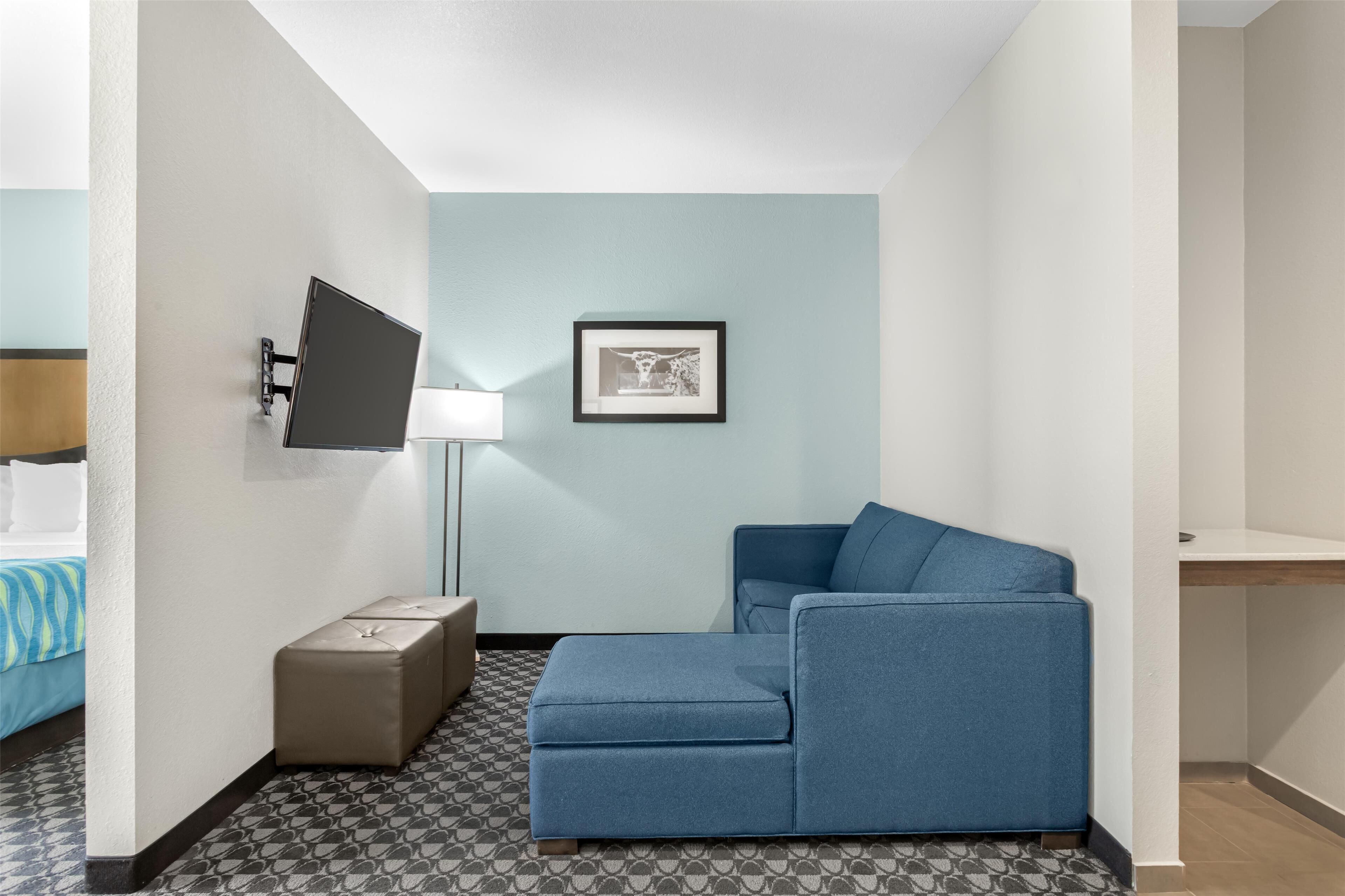 A modern hotel room with a blue sofa, a flat-screen TV mounted on the wall, a bedside table with a lamp, and a framed picture on the wall.
