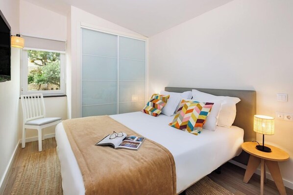 Un dormitorio ordenado con cama, una silla, una mesita de noche con lámpara y una ventana con persianas.