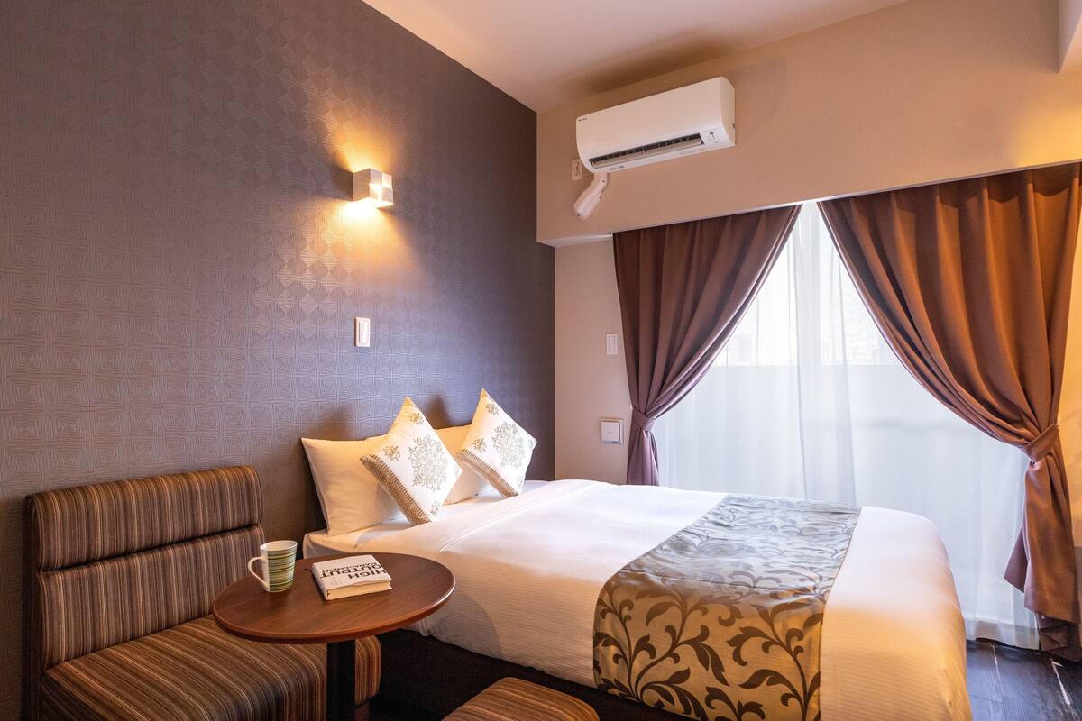 A hotel room with a bed, a seating area, a table, and an air conditioning unit.