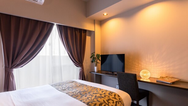 A hotel room with a bed, a desk with a TV, a chair, and a plant. - Mr.KINJO in SAKAEMACHI (Naha)