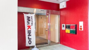 A hotel entrance with a red wall, a glass door, a banner reading "Mr. Kinjo Hotel," and a luggage storage unit with color-coded compartments. - Mr.KINJO in SAKAEMACHI (Naha)