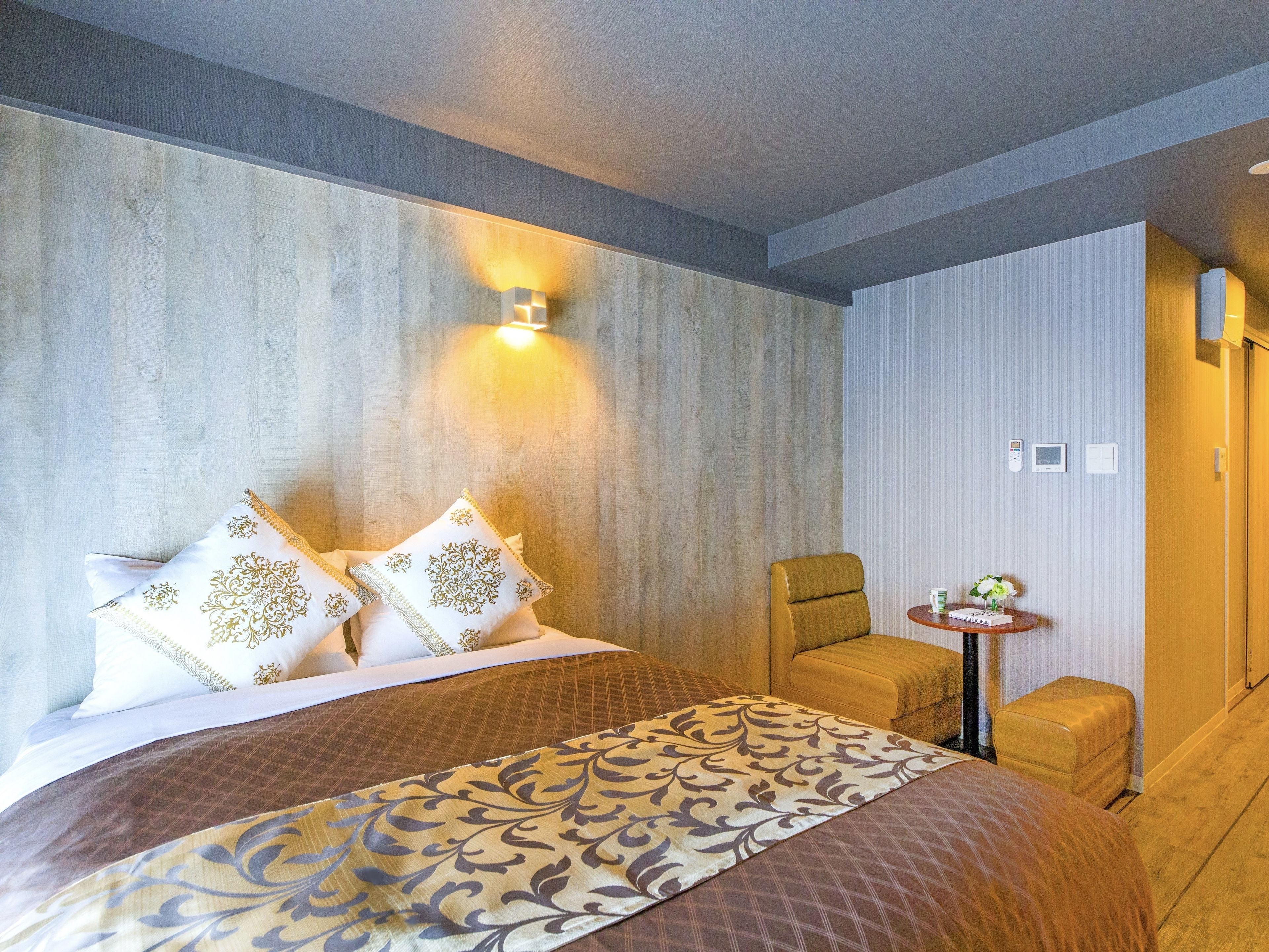 A hotel room with a large bed, a small armchair, a side table with a vase, and a wall-mounted light.