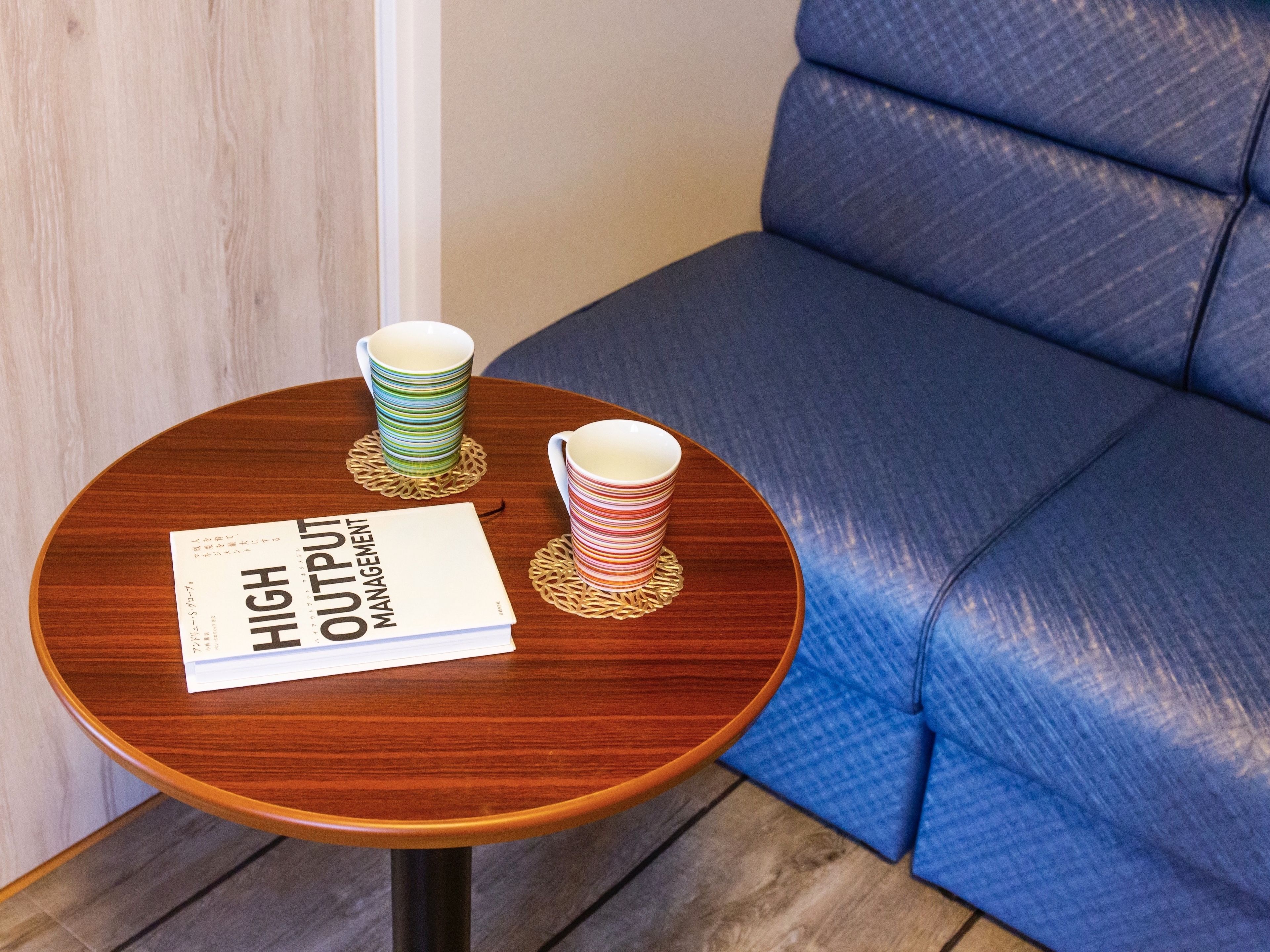 A round wooden table with two stacked cups on coasters, a book titled "HIGH OUTPUT MANAGEMENT," and a blue upholstered chair.
