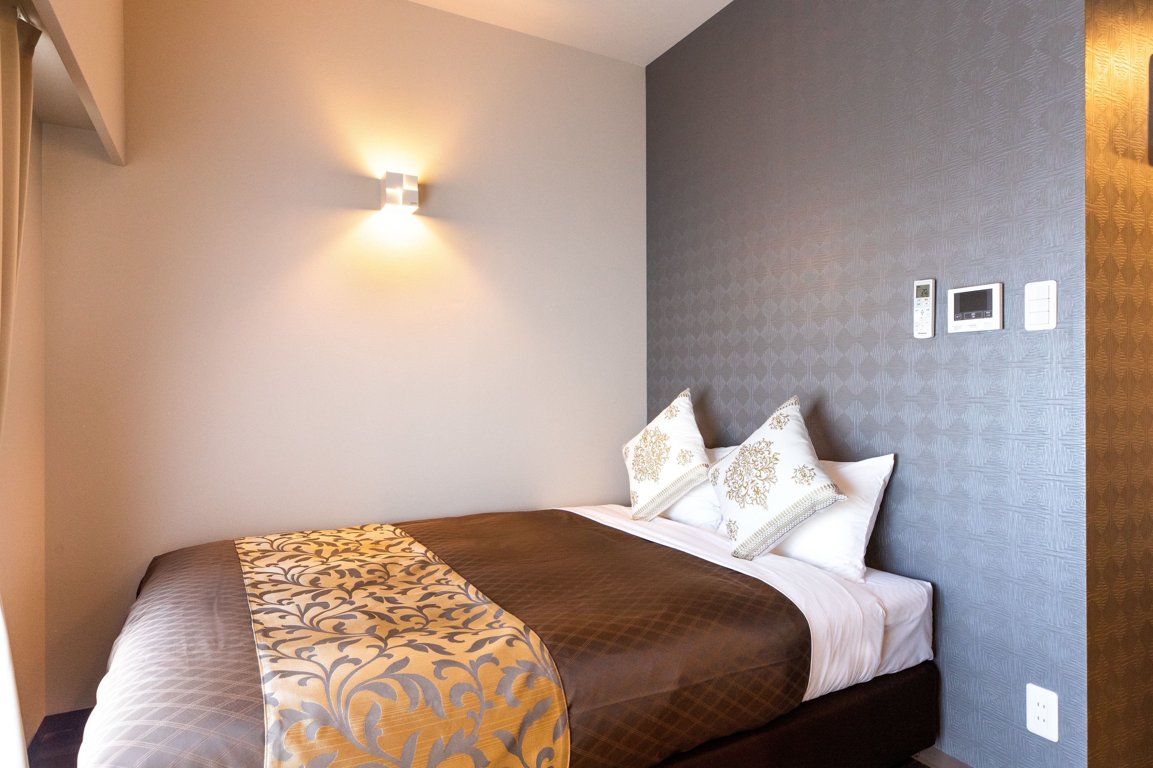 Uma cama bem arrumada com colcha estampada, uma luminária fixada na parede e um termostato na parede.