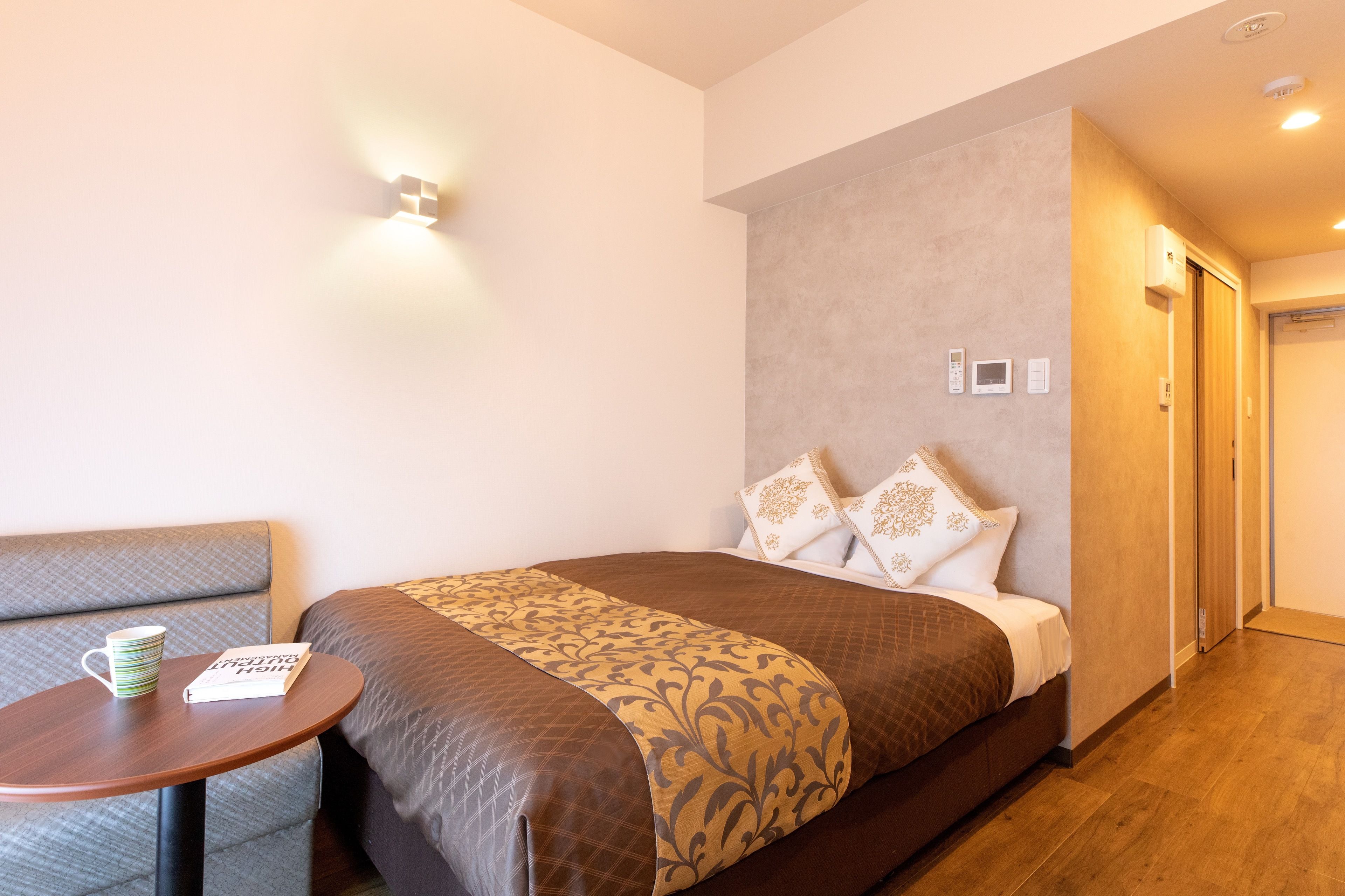 A hotel room with a bed, a small table with a mug and book, a sofa, and a door.