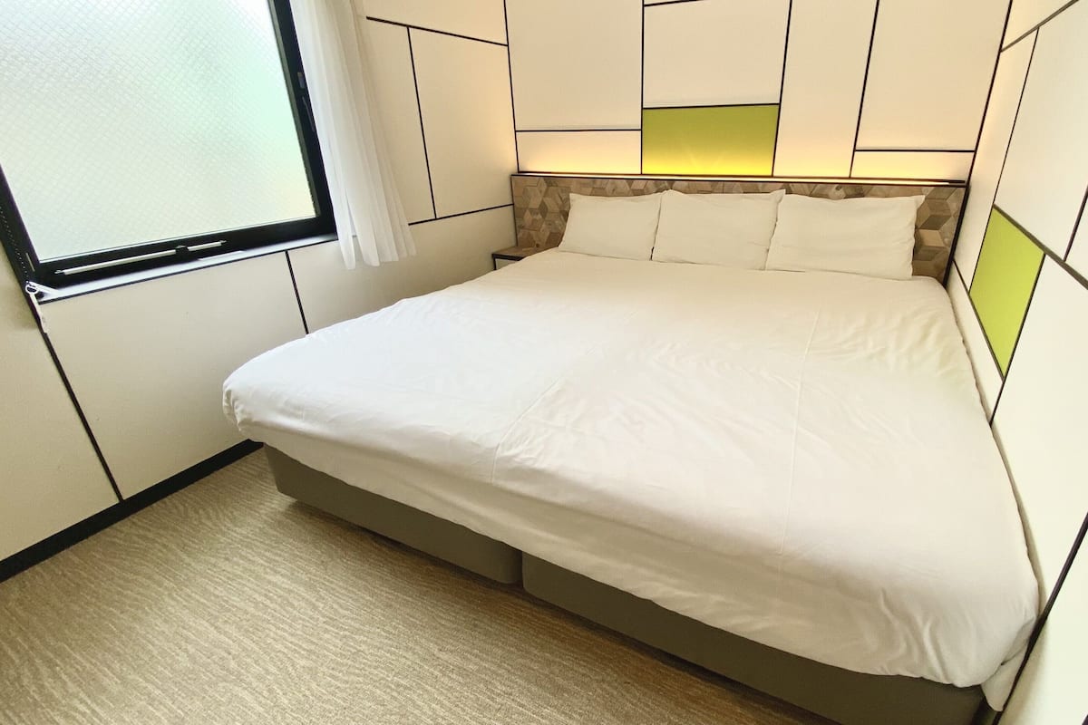 Un lit bien fait dans une chambre dont la tête de lit présente un motif géométrique et qui est dotée d’une fenêtre avec un rideau blanc.