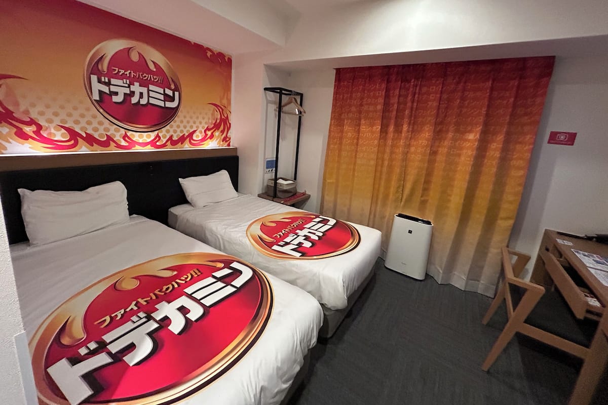Habitación de hotel con dos camas, un escritorio, una silla, una ventana con cortinas y una pared con un logotipo grande.