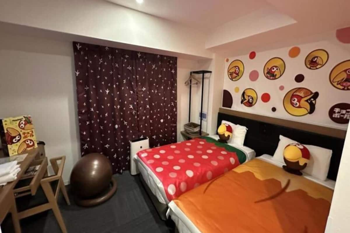 Habitación de hotel con dos camas, un escritorio, una silla, una pelota y una pared decorada con personajes de dibujos animados.