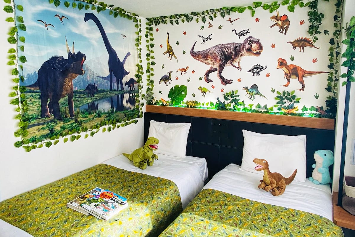 Una habitación con dos camas, decoración de paredes con temática de dinosaurios y adornos con hojas verdes.