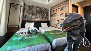 Een hotelkamer met twee bedden, beddengoed met dinosaurusmotieven, een muurschildering en een groot dinosaurusbeeld.