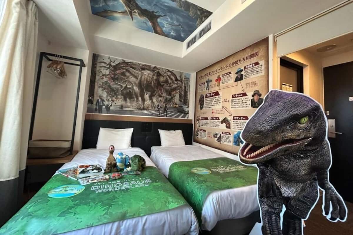 Habitación de hotel con dos camas, ropa de cama con temática de dinosaurios, un mural y una gran figura de dinosaurio.