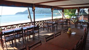 An outdoor dining area with wooden tables and chairs, overlooking a beach and the sea. - Juara Mutiara Resort (Tioman Island)
