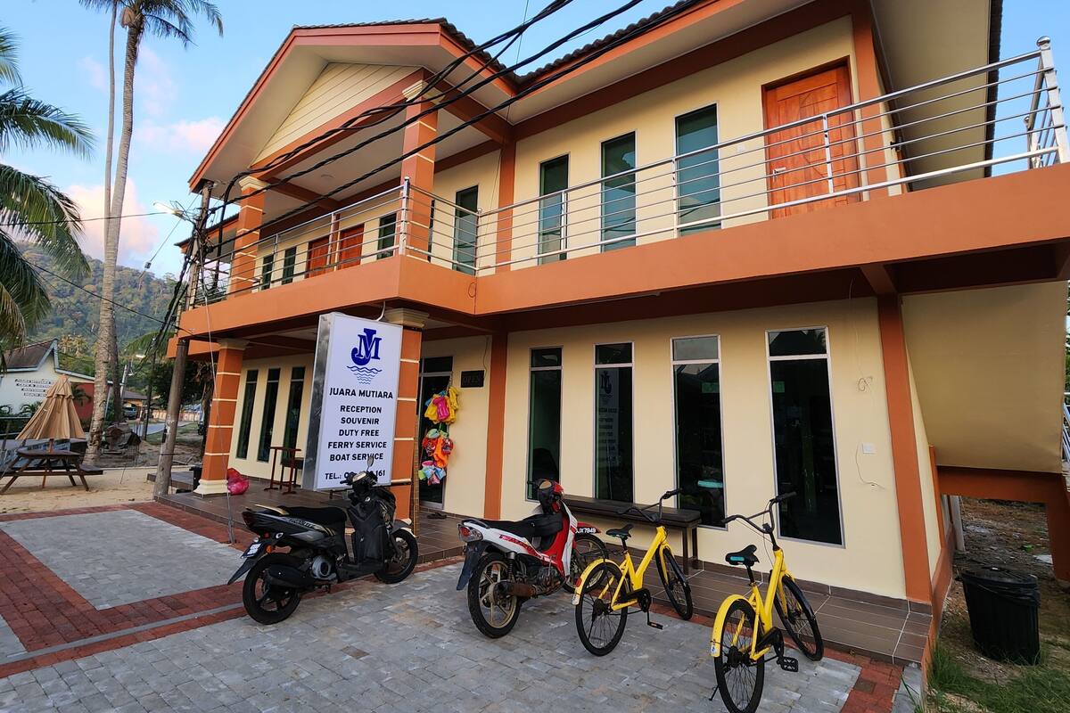 A two-story building with a signboard for 'JUARA MUTIARA' indicating reception, souvenir, duty-free, ferry service, and boat service. There are two bicycles and a scooter parked in front.