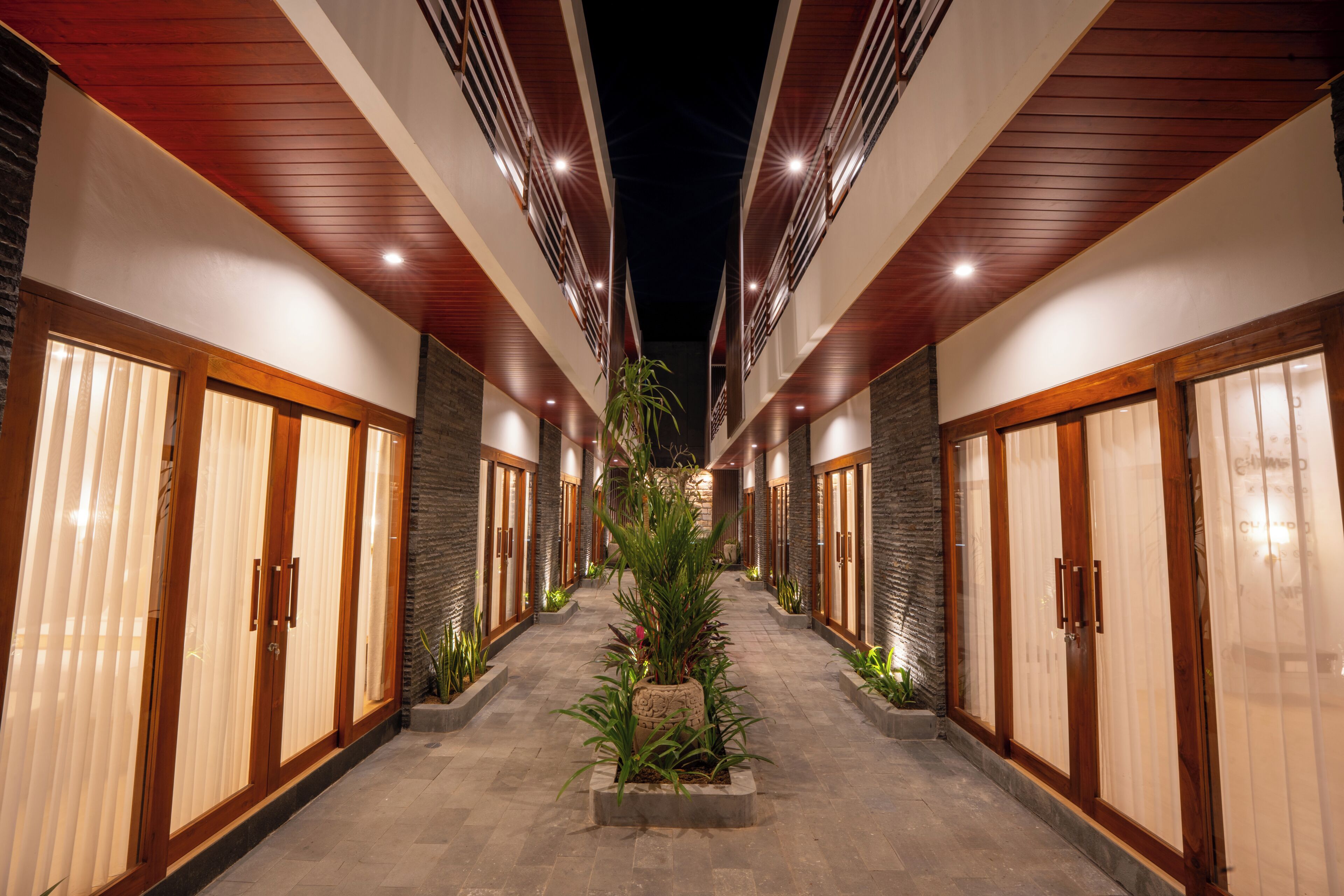 A corridor with wooden doors and stone walls, leading to a central area with plants.