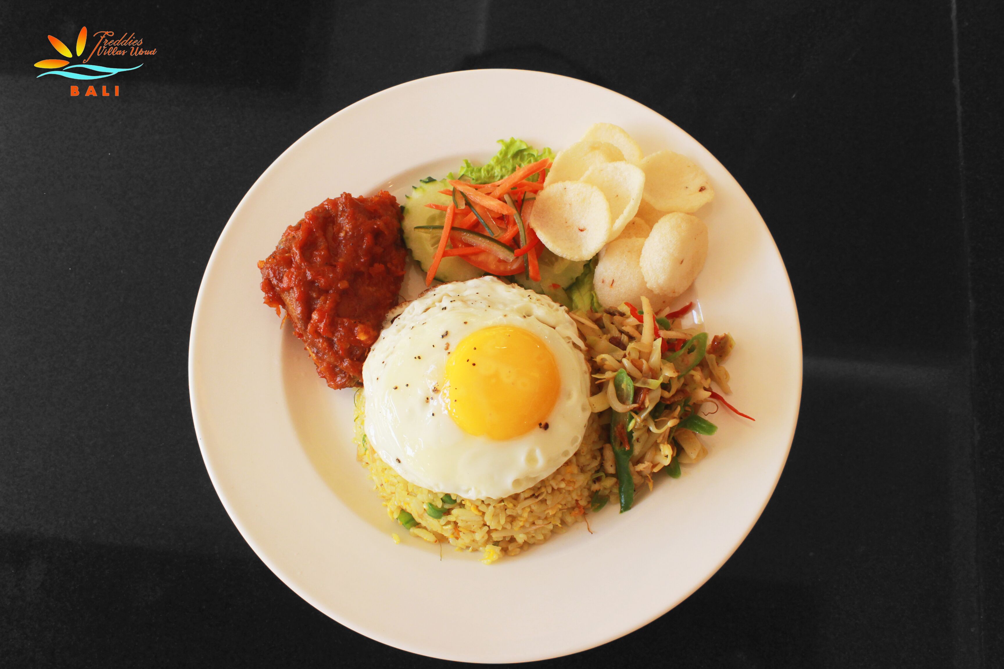 Un plato de comida con un huevo frito, arroz, verduras y una guarnición de sambal y papas fritas.