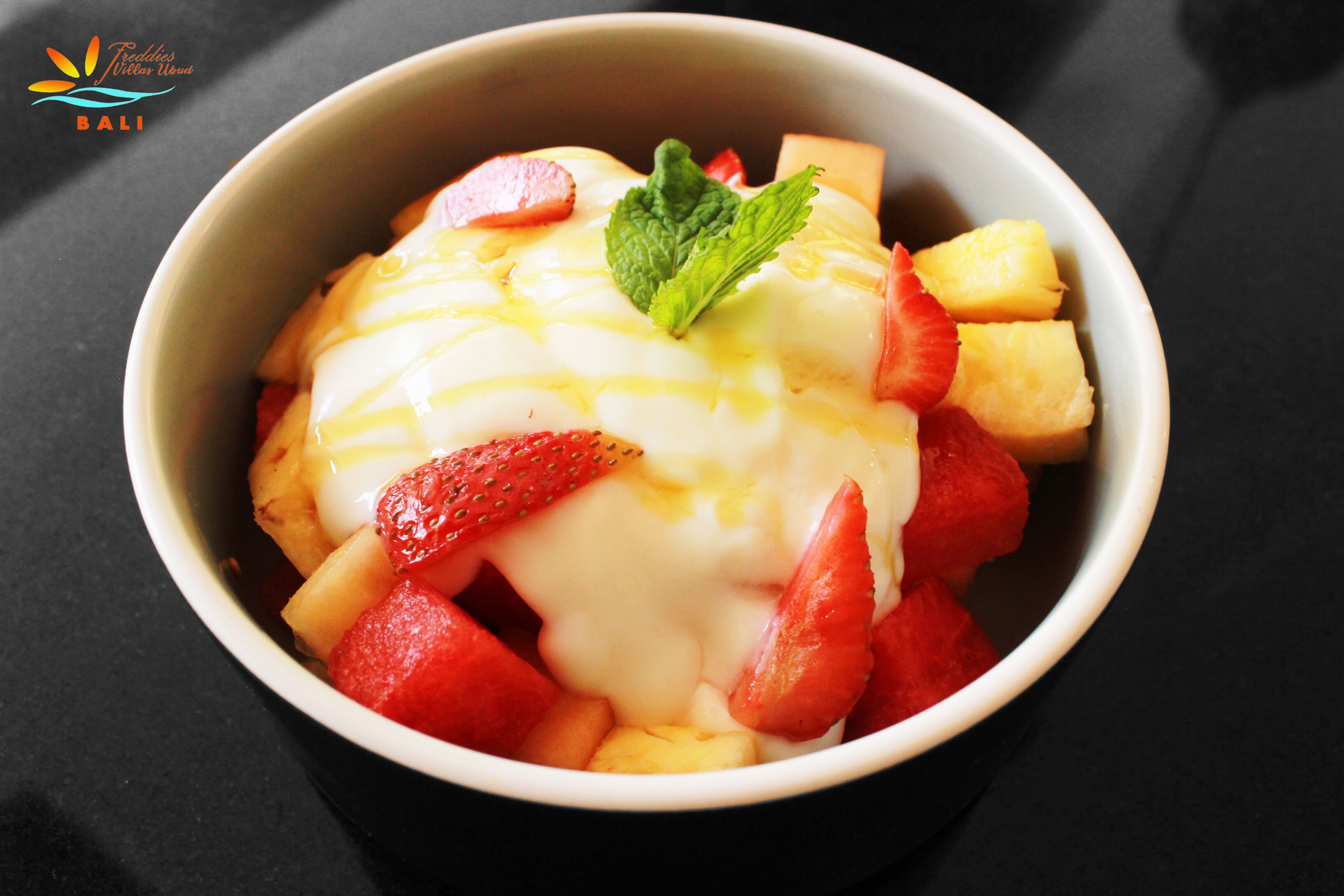 Un tazón de ensalada de frutas con fresas, piña y una ramita de menta.