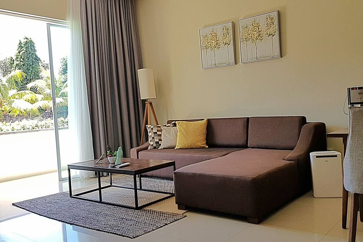 A modern living room with a brown sofa, a coffee table, and a chair. There are two framed artworks on the wall and a window with curtains.