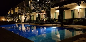 A nighttime pool area with lounge chairs and umbrellas, surrounded by a building with large windows and a well-lit exterior.