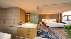 A modern hotel room with a large bed, a separate bathroom, a small seating area, and a view of the city.