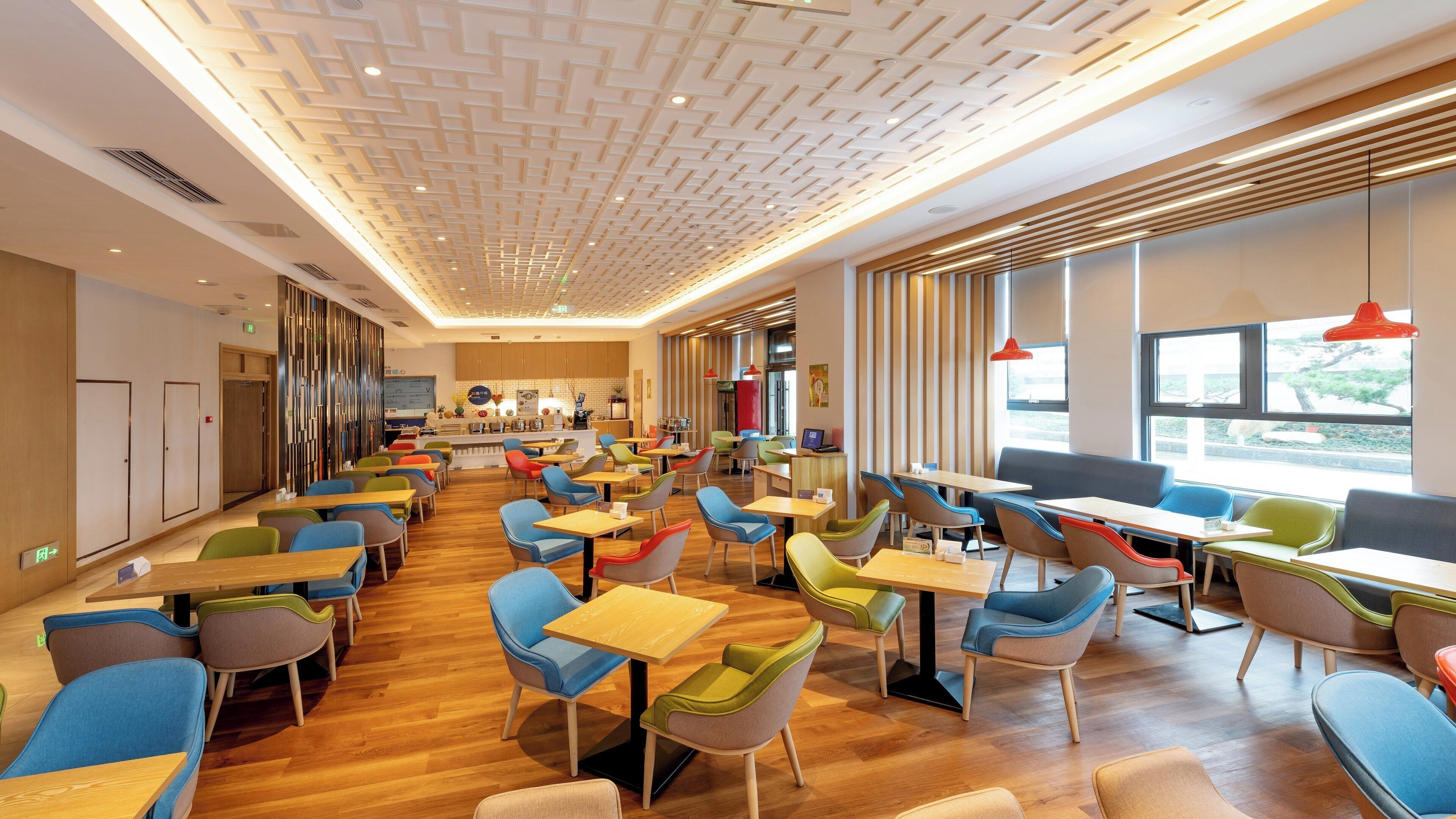 A modern dining area with multiple tables and chairs, a kitchen in the background, and large windows.