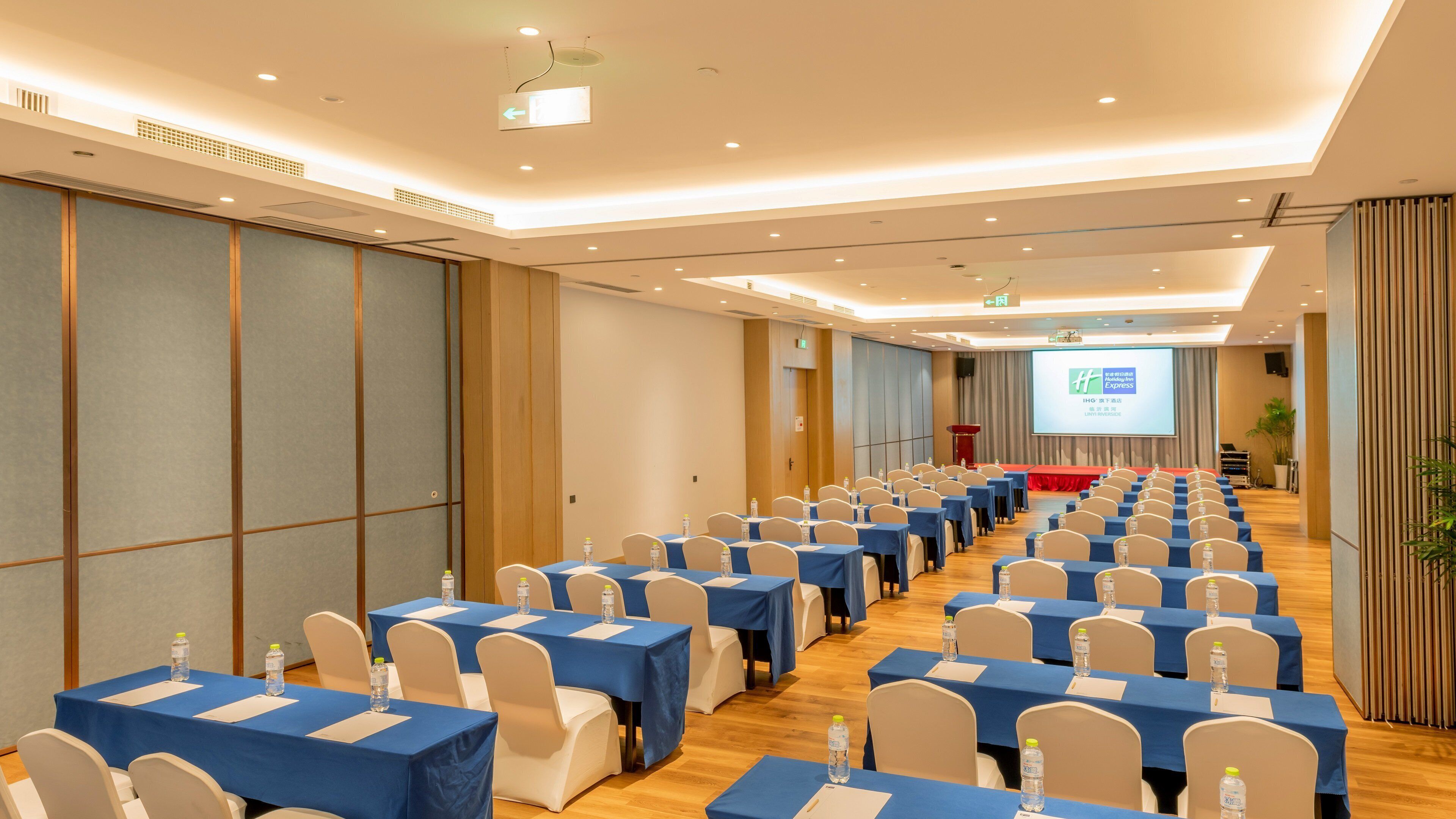 A conference room with rows of tables and chairs, a projector screen, and a stage.